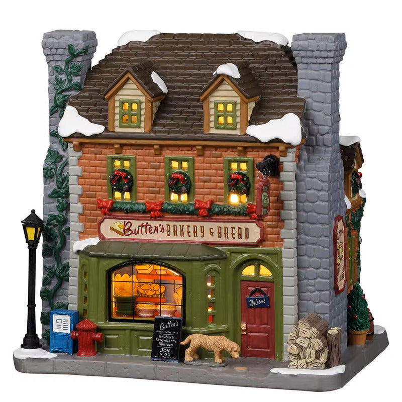 Butter's Bakery & Bread Lemax panificio illuminato villaggio di Natale con vetrina dorata, decorazioni rosse e cane davanti al negozio. | Dematteis.it