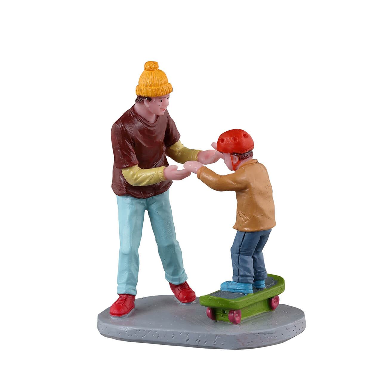 Statuina di un padre che insegna al figlio ad andare sullo skateboard, con entrambe le figure su una piccola piattaforma dettagliata. Il bambino indossa un casco rosso e il padre lo sostiene con le mani tese. La statuina presenta colori vivaci e una scena giocosa, tipica degli accessori per i villaggi Lemax -  Dematteis.it