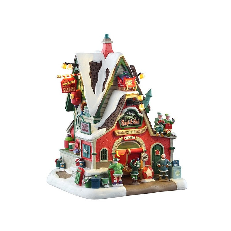 The Elf Service Sleigh & Sled Repair Lemax edificio illuminato del Villaggio Natale con elfi lavoratori, tetto verde innevato e insegne colorate | Dematteis.it
